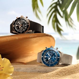 Zwei Breitling Superocean Heritage Chronographen in Schwarz und Blau auf Sand vor tropischer Kulisse mit Palmen.