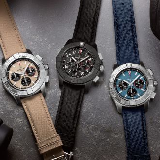 Drei Breitling Avenger Chronographen auf grauem Hintergrund, links in Stahl, beiges Zifferblatt, mittig schwarze Keramik, schwarzes Zifferblatt, rechts in Stahl, blaues Zifferblatt