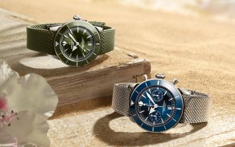Zwei Breitling Superocean Heritage Uhren in Grün und Blau auf Sand und Treibholz vor sommerlicher Kulisse.