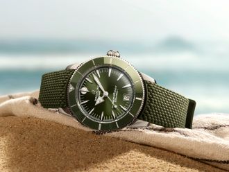 Eine grüne Breitling Superocean Heritage Armbanduhr mit grünem Textilarmband liegt auf einem sandigen Strandtuch, im Hintergrund ist unscharf das Meer mit sanften Wellen zu sehen.