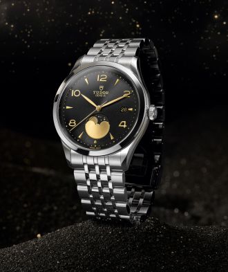 TUDOR 1926 Luna mit schwarzem Zifferblatt, goldener Mondphasenanzeige und Edelstahlarmband auf dunklem Sand mit goldenen Lichtpunkten.
