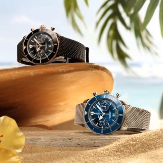 Zwei Breitling Superocean Heritage Chronographen in Schwarz und Blau auf Sand vor tropischer Kulisse mit Palmen.