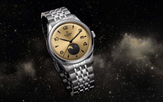 Produktaufnahme der TUDOR 1926 Luna mit champagnerfarbenem Zifferblatt, schwarzer Mondphasenanzeige und Edelstahlarmband vor Sternenhimmel.