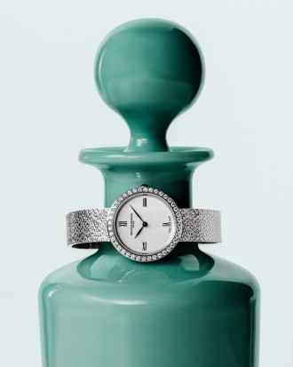 aume & Mercier Joia Luxus-Damenuhr mit Diamantlünette, weißem Zifferblatt und feinem Milanaise-Armband aus Weißgold, stilvoll fotografiert vor einer dekorativen Glasvase.