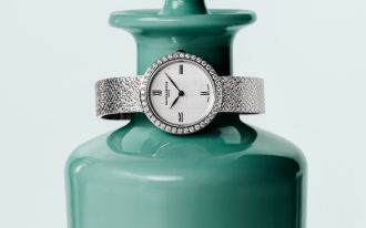 Baume & Mercier Joia Luxus-Damenuhr mit Diamantl&uuml;nette, wei&szlig;em Zifferblatt und feinem Milanaise-Armband aus Wei&szlig;gold, stilvoll fotografiert vor einer dekorativen Glasvase.