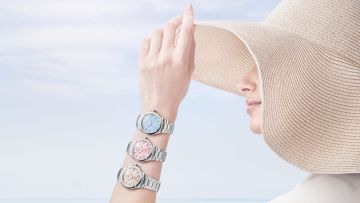 Frau mit großem Sonnenhut am Meer trägt drei Riviera Uhren von Baume & Mercier in Pastellfarben, blau, rosa, beige, am Unterarm.