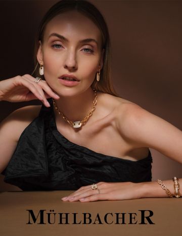 Model trägt exklusiven Goldschmuck von Juwelier Mühlbacher – Goldkette mit diamantbesetztem Anhänger, passende Ohrringe, Ringe und Armband, elegante Inszenierung vor warmem Hintergrund
