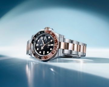 Rolex GMT-Master II in Stahl Rosegold mit schwarzem Zifferblatt und schwarz-brauner Lünette seitlich auf blauem Hintergrund liegend