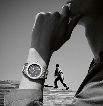 Maurice Lacroix Aikonic Uhr am Handgelenk eines Mannes in Denkerpose, im Hintergrund ein Mann auf Holztreppe