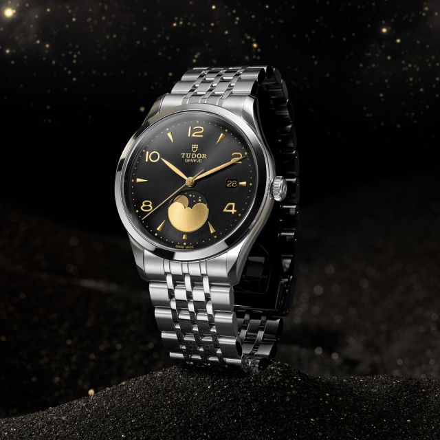 TUDOR 1926 Luna mit schwarzem Zifferblatt, goldener Mondphasenanzeige und Edelstahlarmband auf dunklem Sand mit goldenen Lichtpunkten.