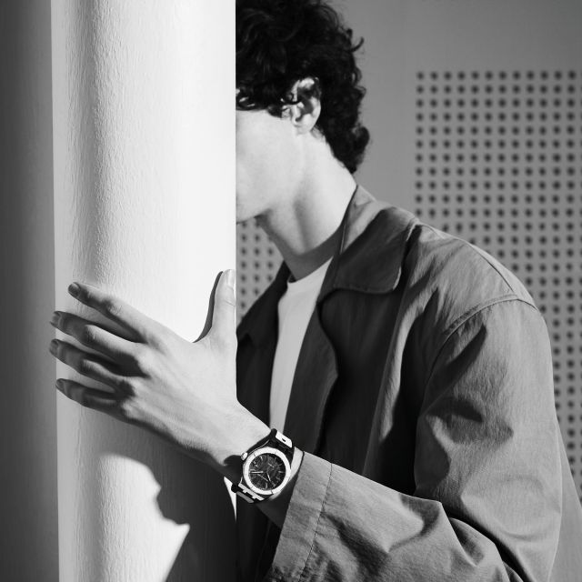 Schwarz-Weiß-Fotografie: Mann lehnt an Säule, Maurice Lacroix AIKONIC Automatikuhr sichtbar, elegante Schweizer Luxusuhren-Szene.