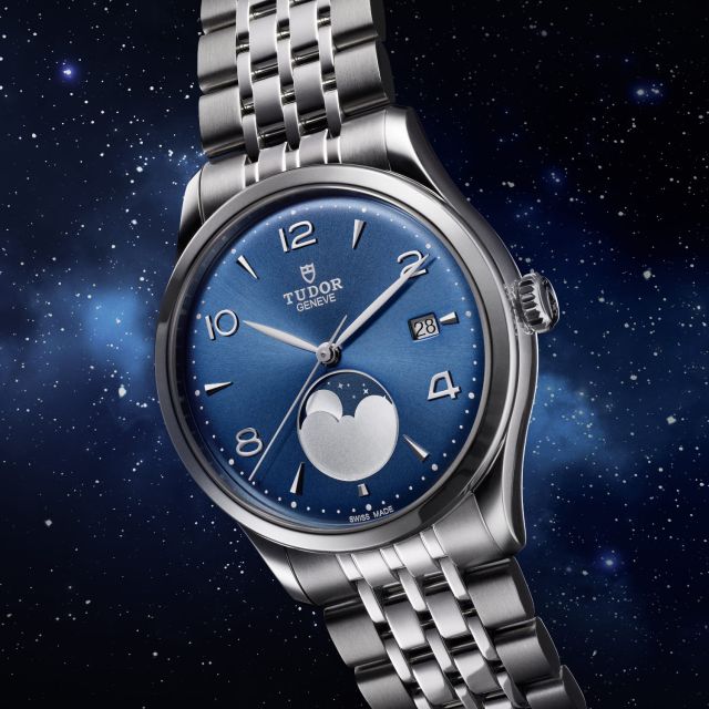 TUDOR 1926 Luna mit blauem Zifferblatt, silberner Mondphasenanzeige und Edelstahlarmband vor nachtblauem Sternenhimmel.