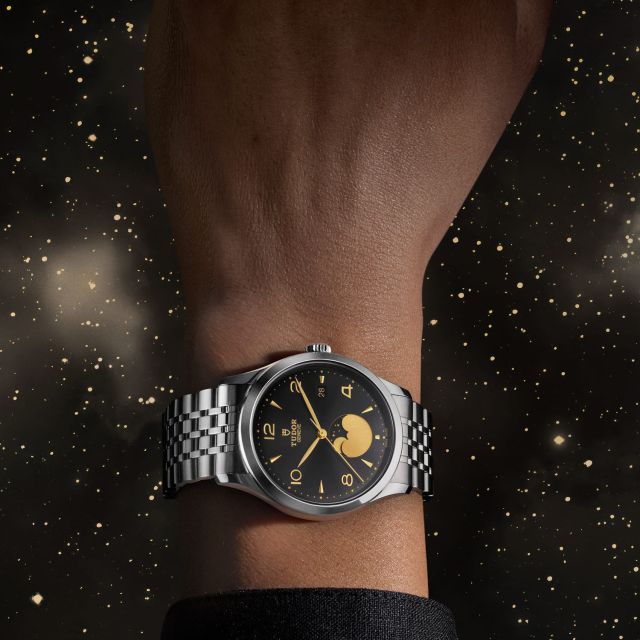 TUDOR 1926 Luna mit schwarzem Zifferblatt und goldener Mondphasenanzeige am Handgelenk vor funkelndem Sternenhimmel.