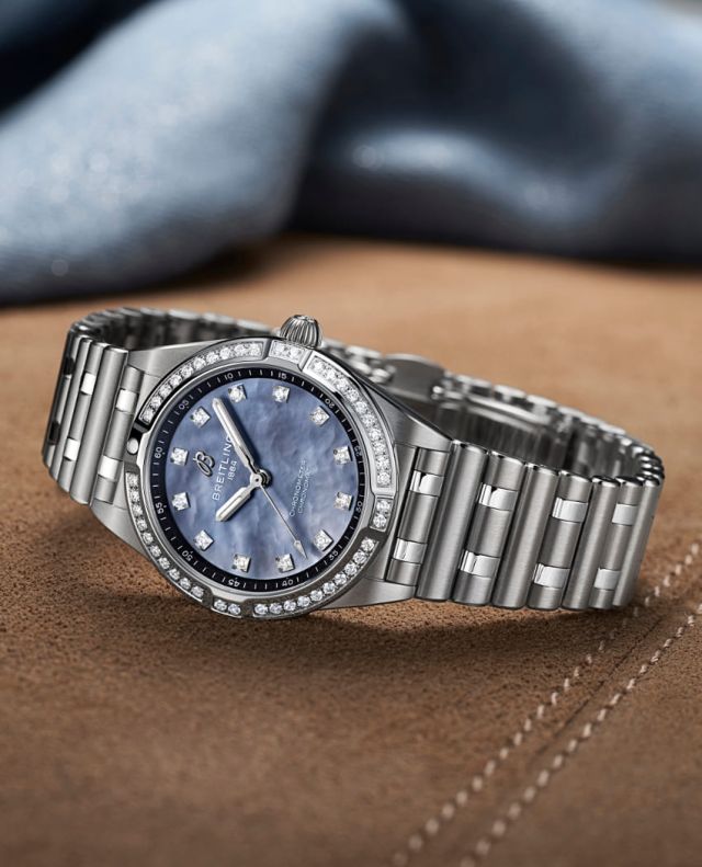 Breitling Chronomat Uhr in Stahl mit blauem Perlmuttzifferblatt und Diamantluenette seitlich auf braunem Textiluntergrund liegend
