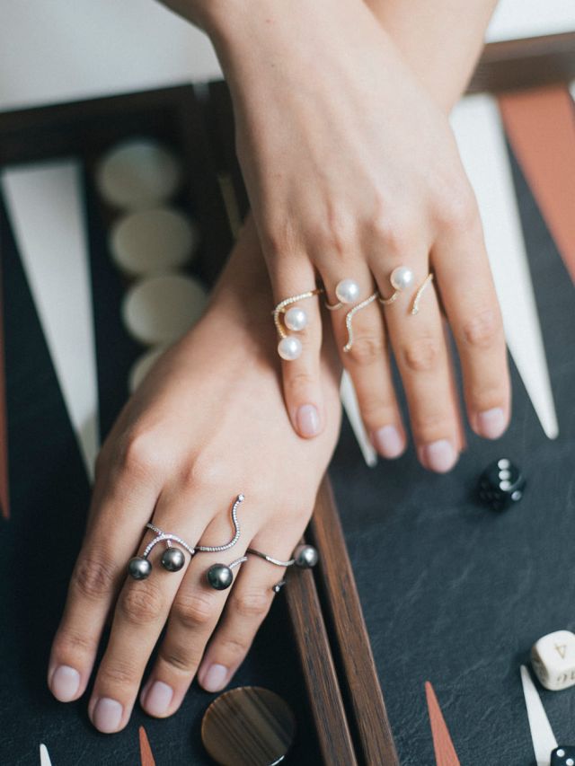 Hände mit modernen Perlenringen aus weißen und dunklen Perlen auf einem Backgammon-Spielbrett, eleganter Schmuck mit filigranem Design.