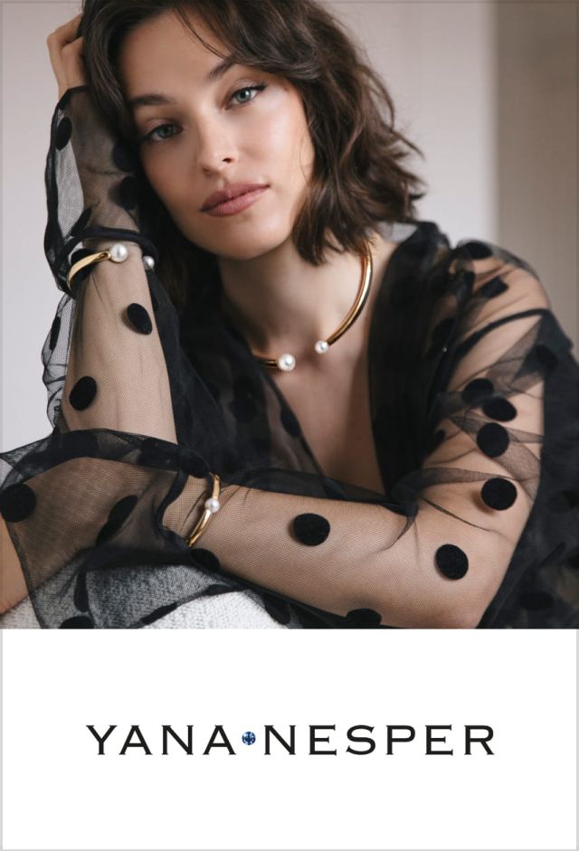 Frau in schwarzer, gepunkteter Tüllbluse trägt goldenen Halsreif und Armreife mit weißen Perlen von Yana Nesper, darunter das Yana Nesper Logo auf weißem Hintergrund.