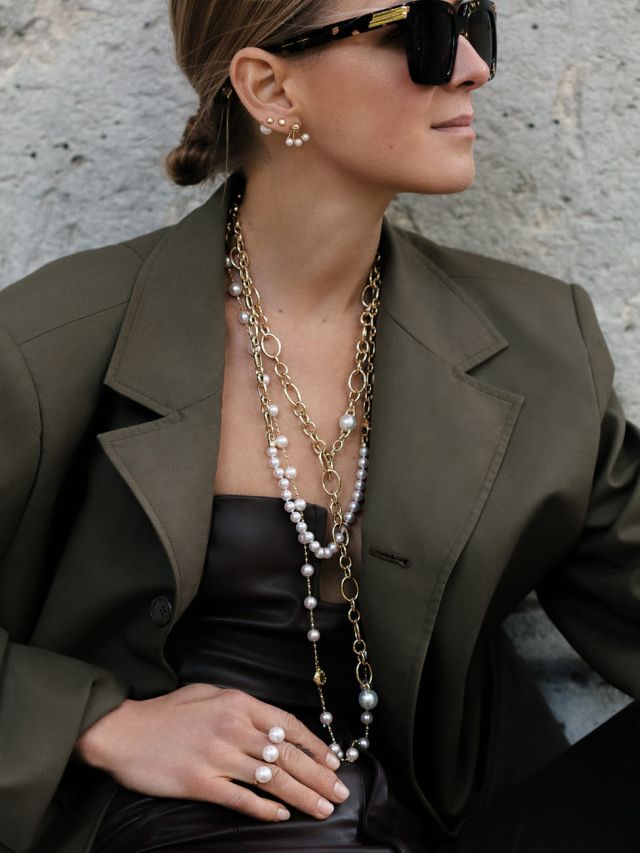 Modebewusste Frau mit Sonnenbrille, langen goldenen Layering-Ketten mit weißen Perlen, Perlenohrringen und olivgrünem Blazer.