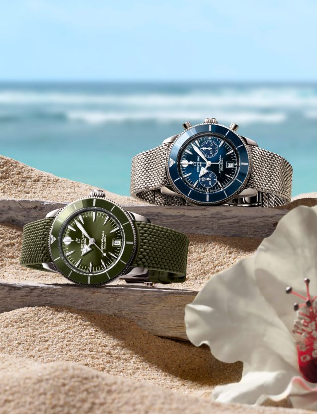 Zwei Breitling Superocean Heritage Uhren in Blau mit Metallarmband und in Grün mit Kautschukarmband liegen dekorativ auf Sand und Treibholz vor tropischem Meereshintergrund.