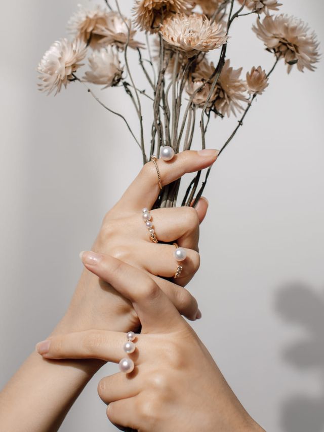 Zarte Hände mit mehreren Perlenringen und Trockenblumen, Schmuck mit weißen Perlen in eleganten Goldfassungen.