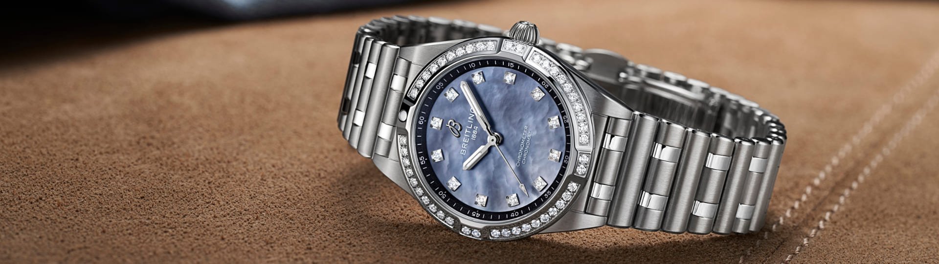 Breitling Chronomat Damenuhr in Stahl mit Diamanluenette, blauem Perlmutzifferblatt und Rouleauxarmband seitlich auf braunem Wildlederbezug liegend