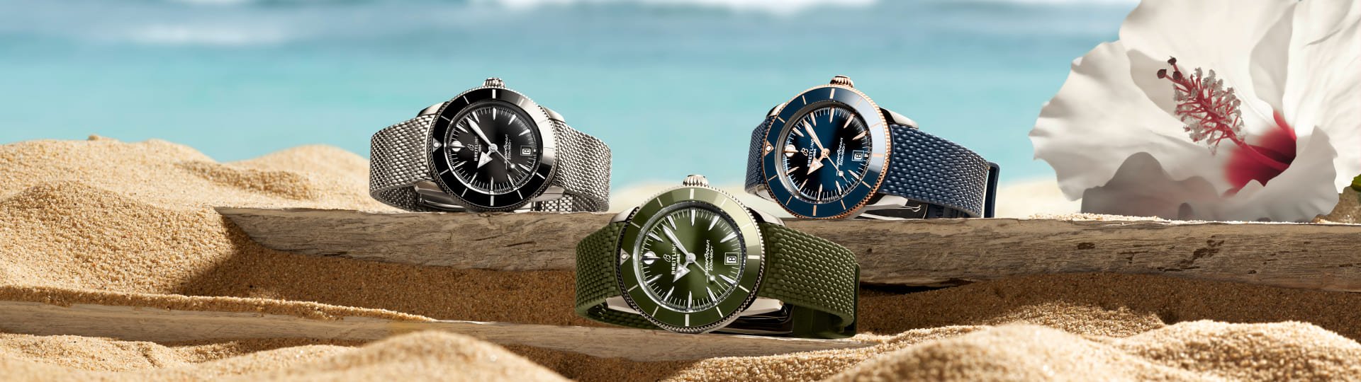 Drei Breitling Superocean Heritage Uhren in Schwarz, Grün und Blau liegen auf Sand und Treibholz vor einem unscharfen Strand- und Meereshintergrund.