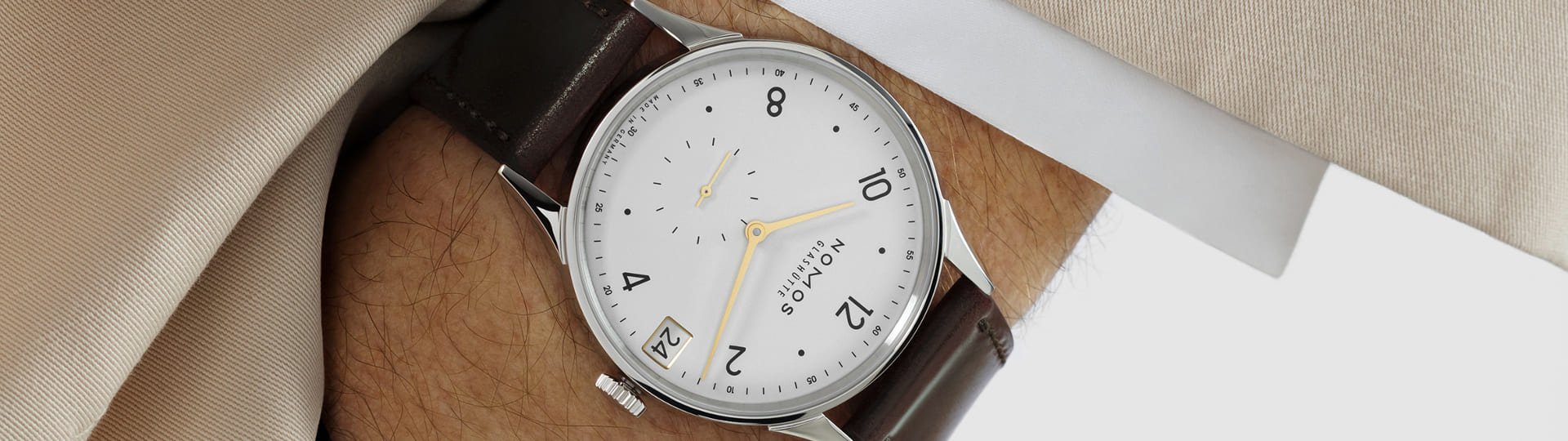 Herrenhandgelenk mit einer Minimatik Uhr von NOMOS Glashuette in Stahl mit weissem Zifferblatt, goldenen Applikationen und einem braunem Lederarmband