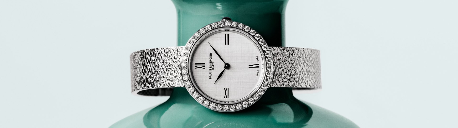 Baume & Mercier Joia Luxus-Damenuhr mit Diamantl&uuml;nette, silbernem Zifferblatt und strukturiertem Metallarmband, stilvoll pr&auml;sentiert vor einer t&uuml;rkisfarbenen Vase.