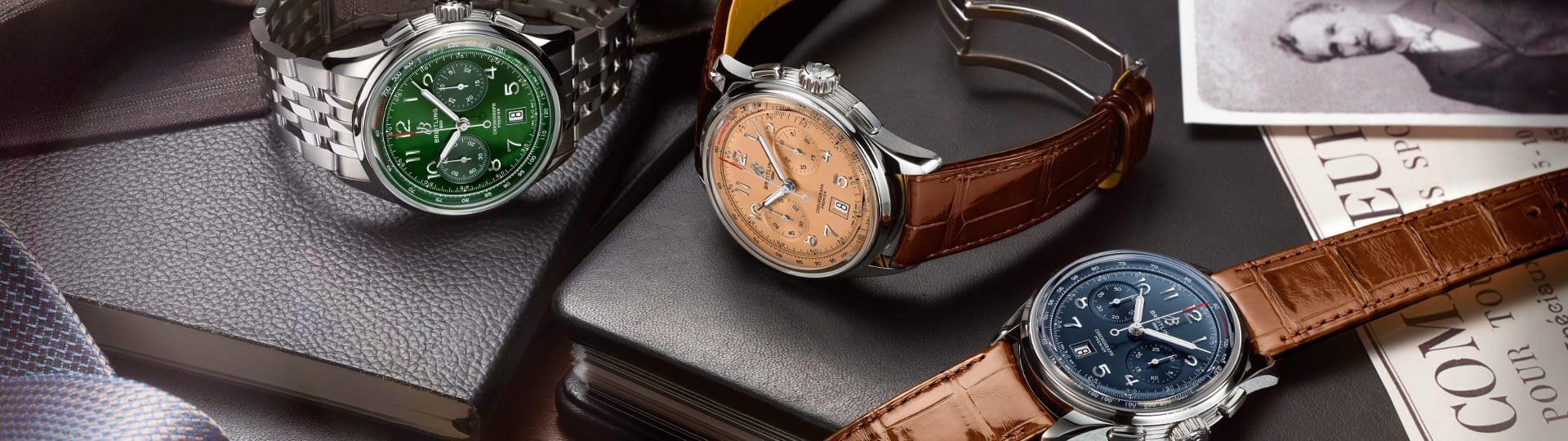 Drei Breitling Premier Chronographen mit Stahlgehaeusen, Zifferblaetter in gruen, kupfer und blau und Armbaender in Stahl und braunem Leder auf Lifestylehintergrund