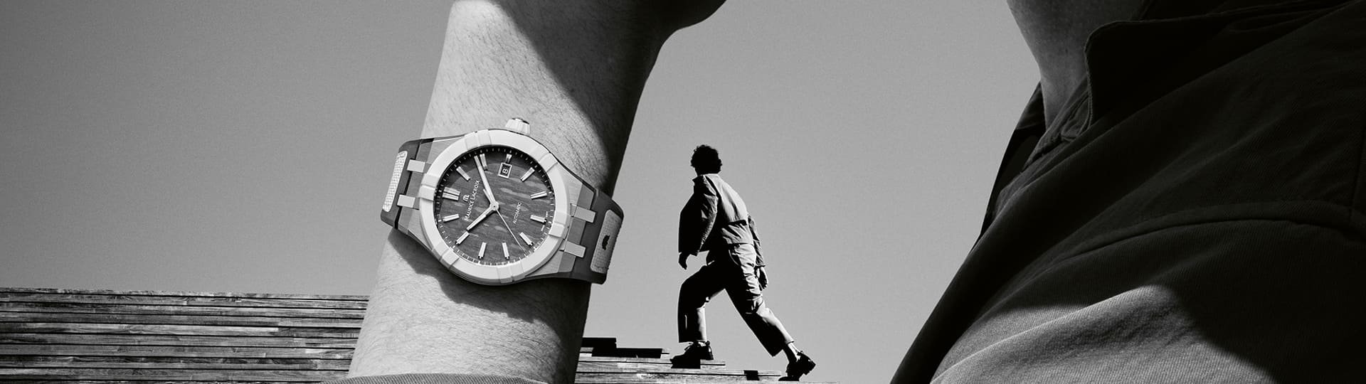 Maurice Lacroix Aikonic Uhr am Handgelenk eines Mannes in Denkerpose, im Hintergrund ein Mann auf Holztreppe