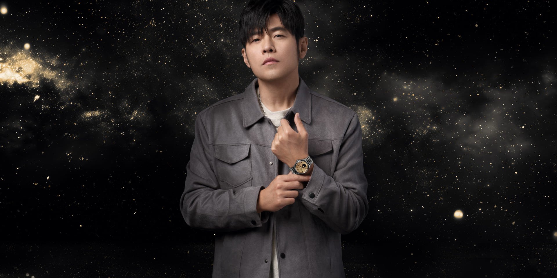 Jay Chou trägt eine TUDOR 1926 Luna Uhr mit champagnerfarbenem Zifferblatt und Mondphasenanzeige vor einem Hintergrund mit funkelndem Sternenhimmel.