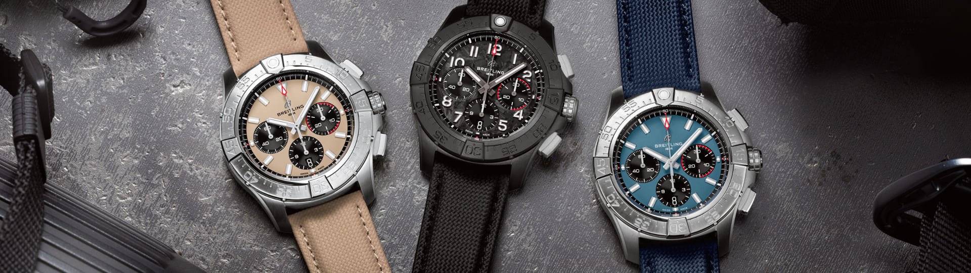 Drei Breitling Avenger Chronographen auf grauem Hintergrund, links in Stahl, beiges Zifferblatt, mittig schwarze Keramik, schwarzes Zifferblatt, rechts in Stahl, blaues Zifferblatt