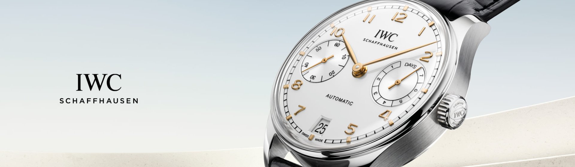IWC Schaffhausen Portugieser Automatic 42 in Stahl mit silberweissem Zifferblatt mit rosegoldenen Applikationen leicht seitlich vor hellblau-beigefarbenem Hintergrund und IWC Logo daneben