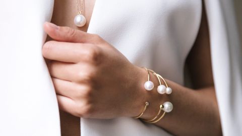 Nahaufnahme einer Hand mit elegantem Armschmuck von Yana Nesper: zarte goldene Armreifen mit weißen Perlen, kombiniert mit einer passenden Perlenkette, minimalistischer und luxuriöser Look.