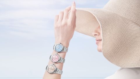 Frau mit großem Sonnenhut am Meer trägt drei Riviera Uhren von Baume & Mercier in Pastellfarben, blau, rosa, beige, am Unterarm.