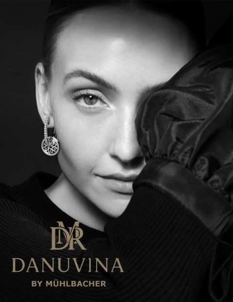 Nahaufnahme einer Frau mit edlen Danuvina Ohrringen by M&uuml;hlbacher, schwarz-wei&szlig; fotografiert, kombiniert mit dem goldfarbenen Markenlogo vor dunklem Hintergrund.