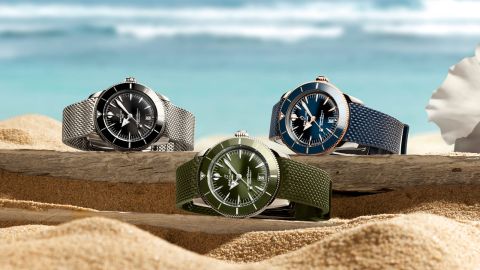 Drei Breitling Superocean Heritage Uhren in Schwarz, Grün und Blau liegen auf Sand und Treibholz vor einem unscharfen Strand- und Meereshintergrund.