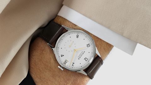 Herrenhandgelenk mit einer Minimatik Uhr von NOMOS Glashuette in Stahl mit weissem Zifferblatt, goldenen Applikationen und einem braunem Lederarmband Herrenhandgelenk mit einer Minimatik Uhr von NOMOS Glashuette in Stahl mit weissem Zifferblatt, goldenen Applikationen und einem braunem Lederarmband