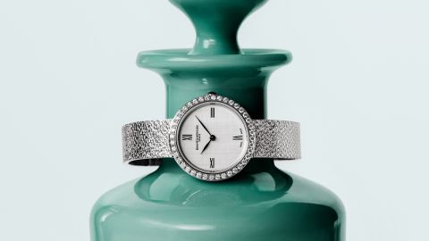 Baume & Mercier Joia Luxus-Damenuhr mit Diamantl&uuml;nette, silbernem Zifferblatt und strukturiertem Metallarmband, stilvoll pr&auml;sentiert vor einer t&uuml;rkisfarbenen Vase.