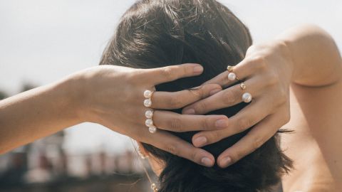 Luxuriöse Perlenringe von Yana Nesper an Frauenhänden, eleganter Goldschmuck mit weißen Süßwasserperlen, stilvolle Nahaufnahme vor hellem Hintergrund.
