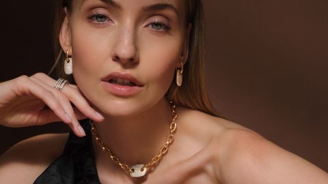 Nahaufnahme einer Frau mit goldener Crivelli-Halskette mit pavébesetztem Anhänger, passenden Ohrringen und diamantbesetzten Ringen an der linken Hand.