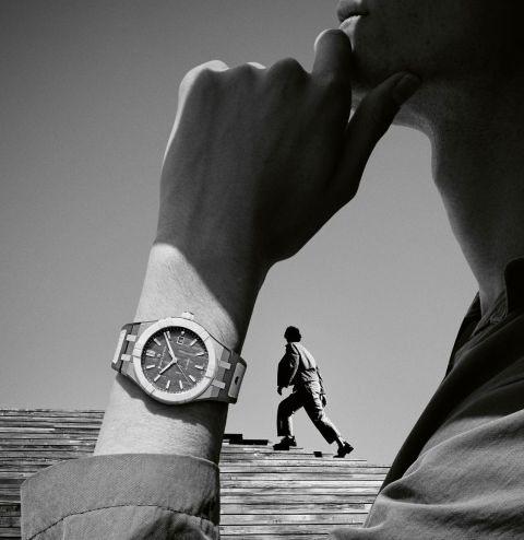 Schwarz-weiß-Werbemotiv mit Maurice Lacroix Aikonic Automatikuhr am Handgelenk, im Hintergrund ein Mann auf Holztreppe Schwarz-weiß-Werbemotiv mit Maurice Lacroix Aikonic Automatikuhr am Handgelenk, im Hintergrund ein Mann auf Holztreppe
