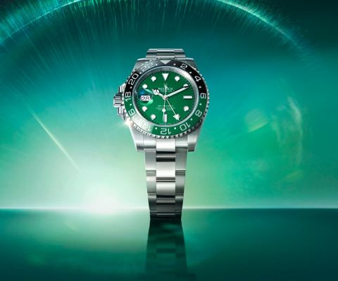 Panoramaaufnahme der Rolex GMT-Master II mit grünem Zifferblatt und schwarzer Cerachrom-Lünette steht im Mittelpunkt auf smaragdgrünem Hintergrund mit Lichteffekt.