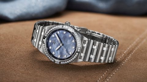 Breitling Chronomat 28 Uhr in Stahl mit Diamantluenette, Rouleauxarmband und Zifferblatt aus blauem Perlmutt seitlich auf braunem Textiluntergrund liegend Breitling Chronomat 28 Uhr in Stahl mit Diamantluenette, Rouleauxarmband und Zifferblatt aus blauem Perlmutt seitlich auf braunem Textiluntergrund liegend