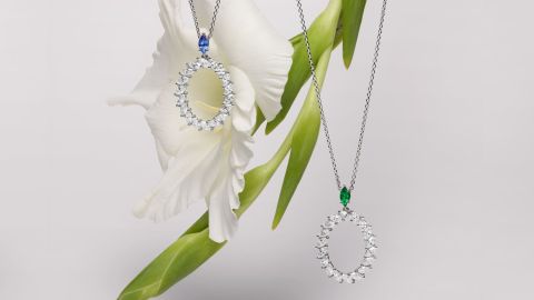 Zwei ovale L'Heure de Diamant Anhaenger mit Diamanten, links mit blauem Saphir, rechts mit Smaragd, je an einer Halskette haengend vor grauem Hintergrund mit weisser Lilie Zwei ovale L'Heure de Diamant Anhaenger mit Diamanten, links mit blauem Saphir, rechts mit Smaragd, je an einer Halskette haengend vor grauem Hintergrund mit weisser Lilie
